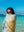 A woman wrapped in a towel with a bold sun design stands by the ocean, gazing at the camera while wearing round sunglasses. The towel features yellow and blue colors against a neutral background, adding a pop of brightness. The crystal-clear turquoise water and deep blue sky in the background create a perfect sunny beach day atmosphere. Her relaxed and playful expression, combined with the vibrant setting, evokes a carefree summer vibe.