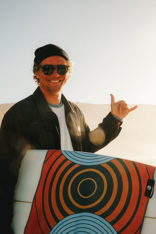 A man on a beach throws up the Shaka Sign, a Happy Faced Towel is draped over his surfboard. Happy and Smiling